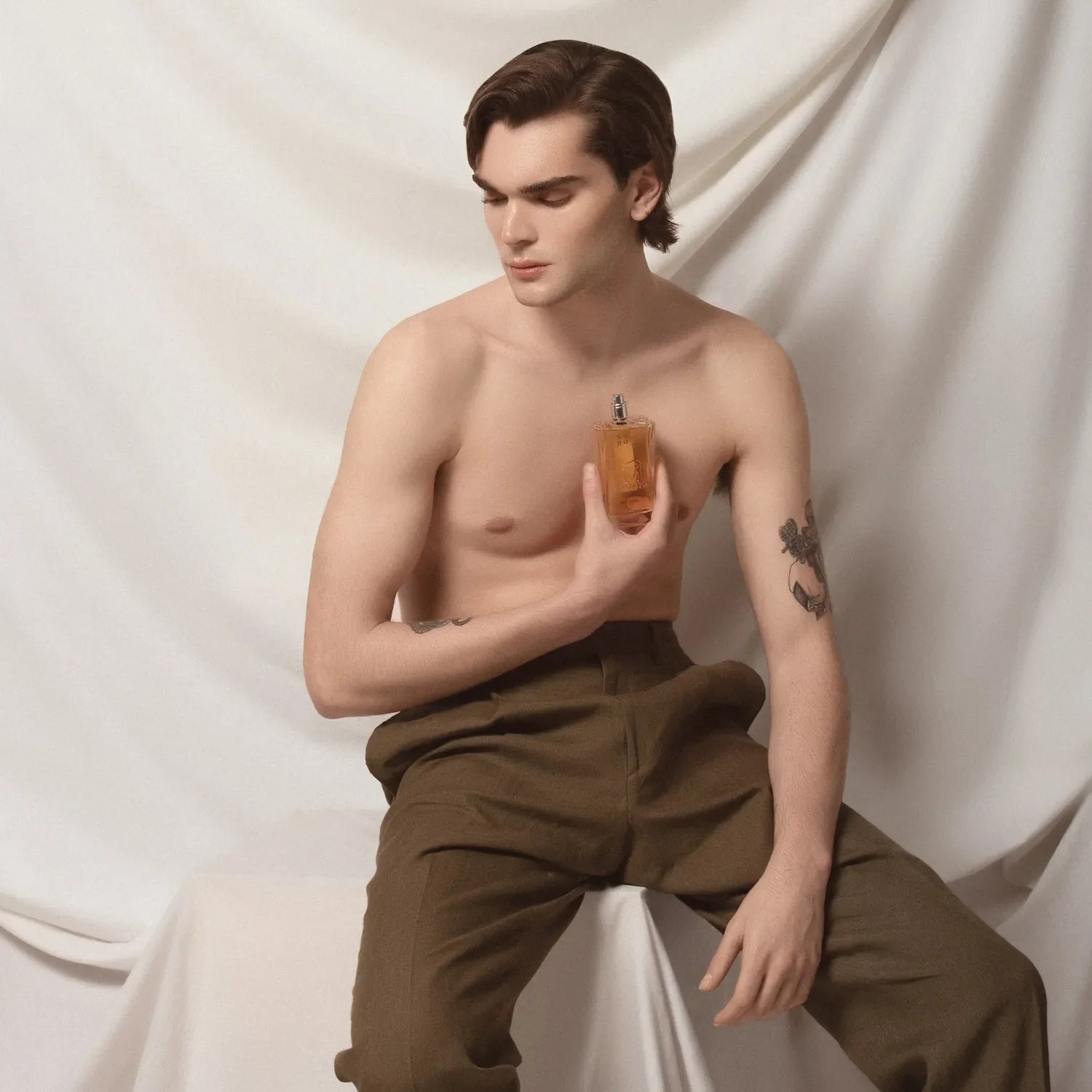 MAn met ontbloot bovenlijk met een fles parfum in zijn linker hand en een tatoeage op zijn rechter bovenarme