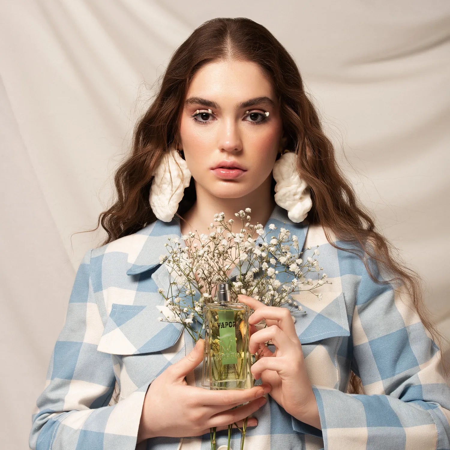 Vrouw met lang haar en een geruit jasje aan houdt bloemen en een fles parfum vast 