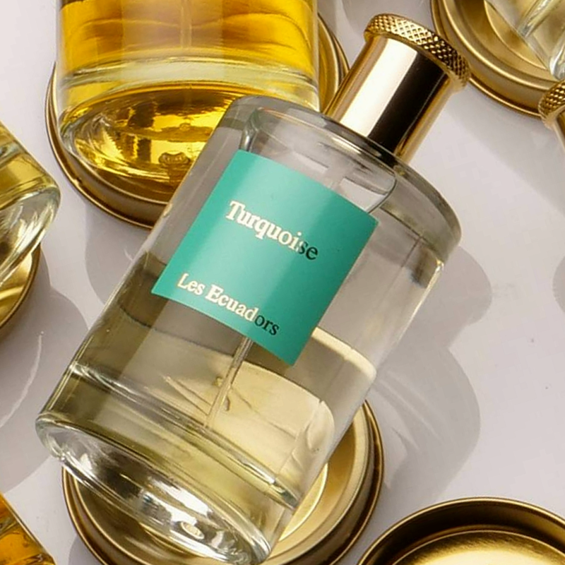 liggende parfumflessen op metalen goudkleurige schijven. de middelste fles heeft een blauw/groen label met de gouden tekst |Turquoise Les Ecuadors